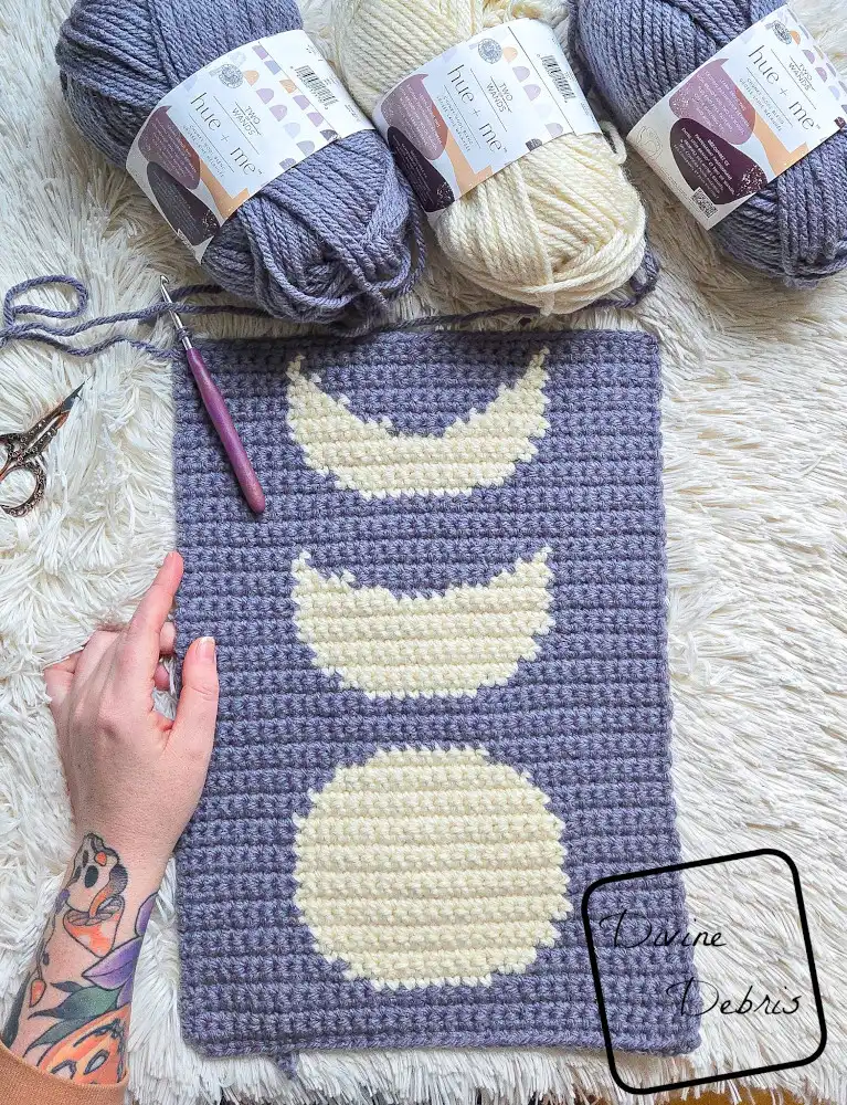 [Image description] Top down look at an unfinished version of the Simple Moon Phase Wall Hanging with skeins of yarn along the top of the photo and a white woman's hand holding the bottom left corner. 