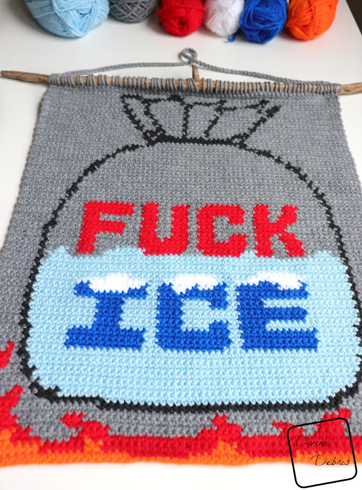 [Image description] A low angle photo of the Fuck Ice Wall Hanging laying on a white background with skeins of yarn along the top of the .photo