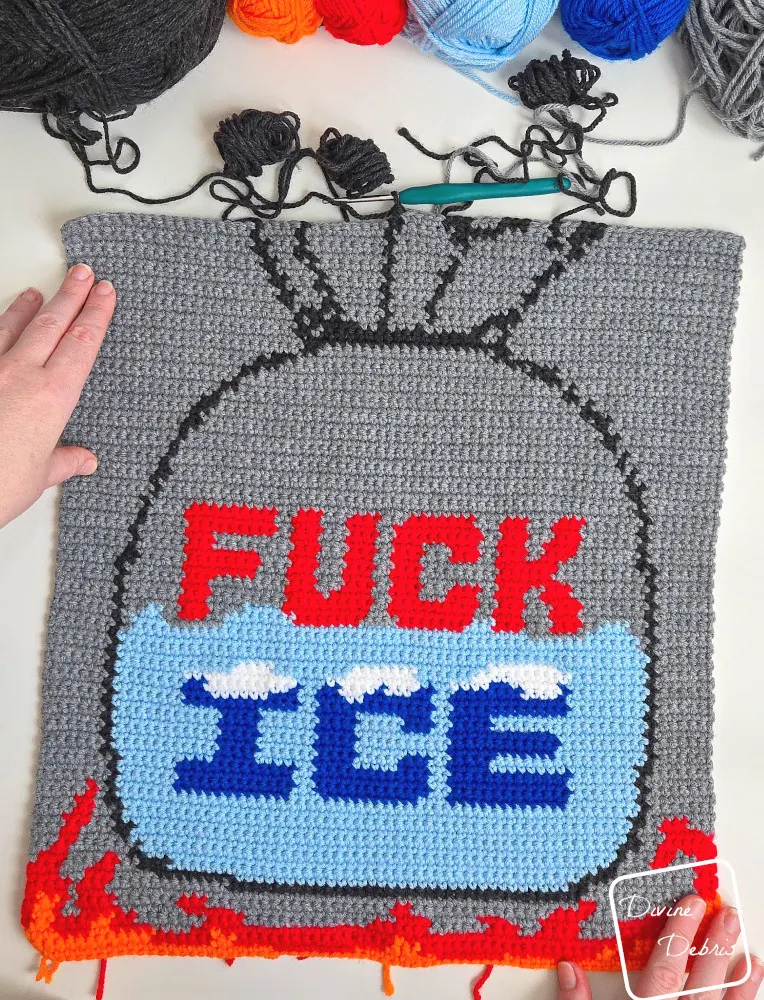 [image description] Top down look at an unfinished Fuck Ice Wall Hanging on a white background with skeins of yarn in the top of the photo and a white woman's hand holding the top left corner and bottom right corner.
