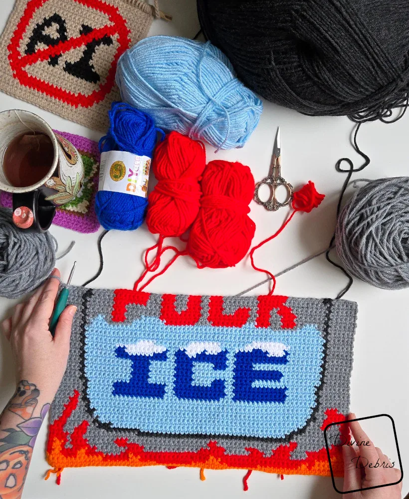 [image description] Top down look at an unfinished Fuck Ice Wall Hanging on a white background with skeins of yarn in the top of the photo and a white woman's hand holding the top left corner and bottom right corner.