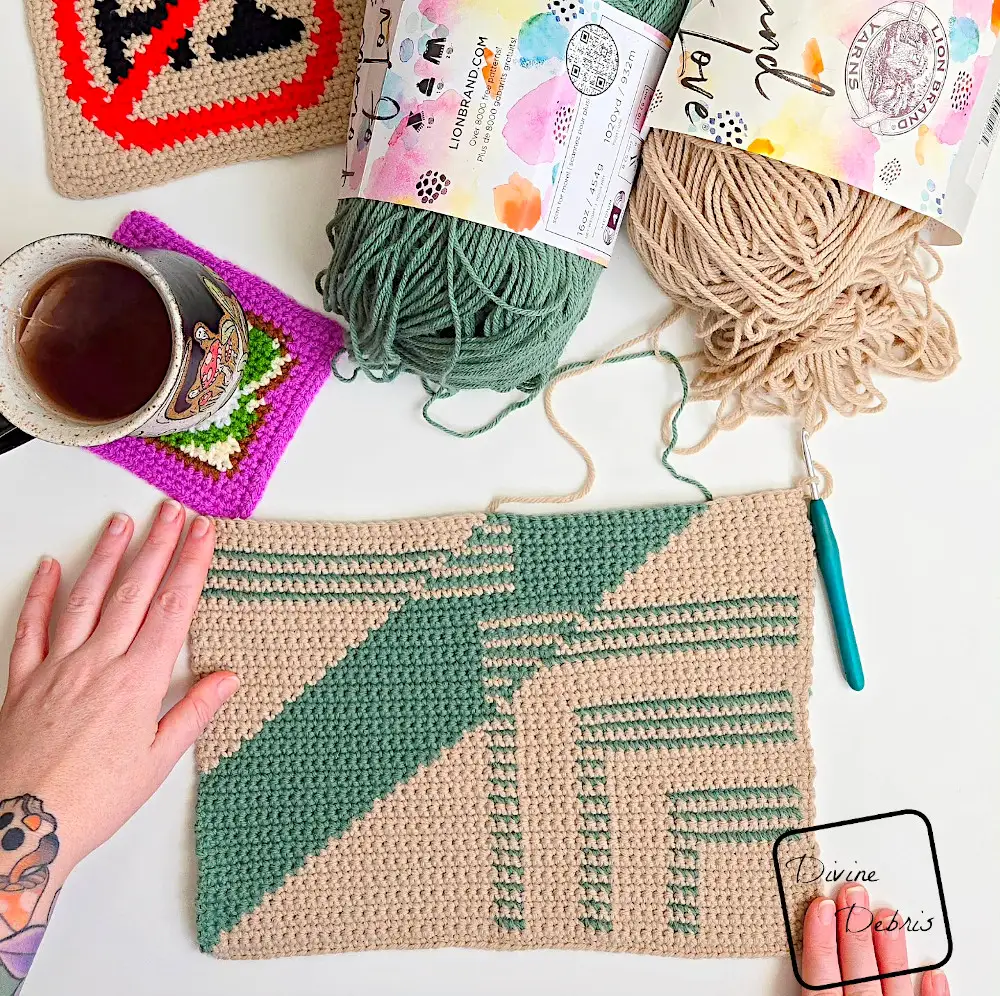 [image description] Top down look at an unfinished Dumpster Fire Wall Hanging on a white background with skeins of yarn in the top of the photo and a white woman's hand holding top left-hand corner and bottom right-hand corner.
