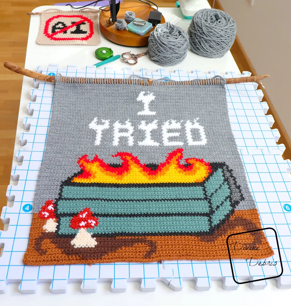 [Image description] A photo of the I Tried Wall Hanging on a blocking mat with cakes of yarn along the top of the photo and crochet tools.