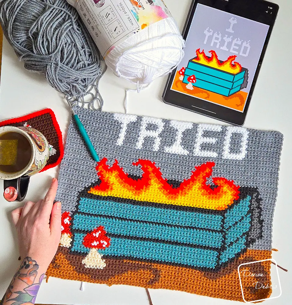 [image description] Top down look at an unfinished Dumpster Fire Wall Hanging on a white background with skeins of yarn in the top of the photo and a white woman's hand holding the bottom left corner.