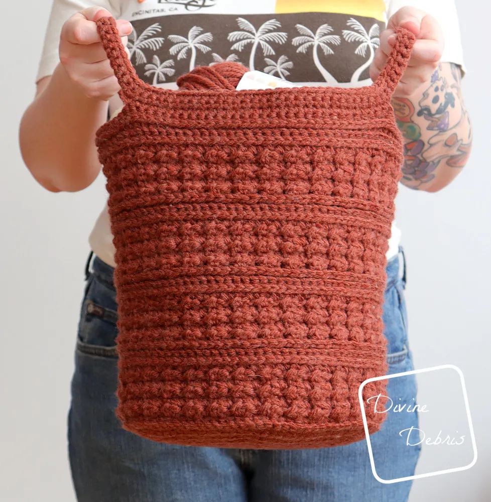 [Image description] A white woman, seen from the shoulders down, stands against a white wall holding the Lucy Basket crochet pattern out toward the viewer. 